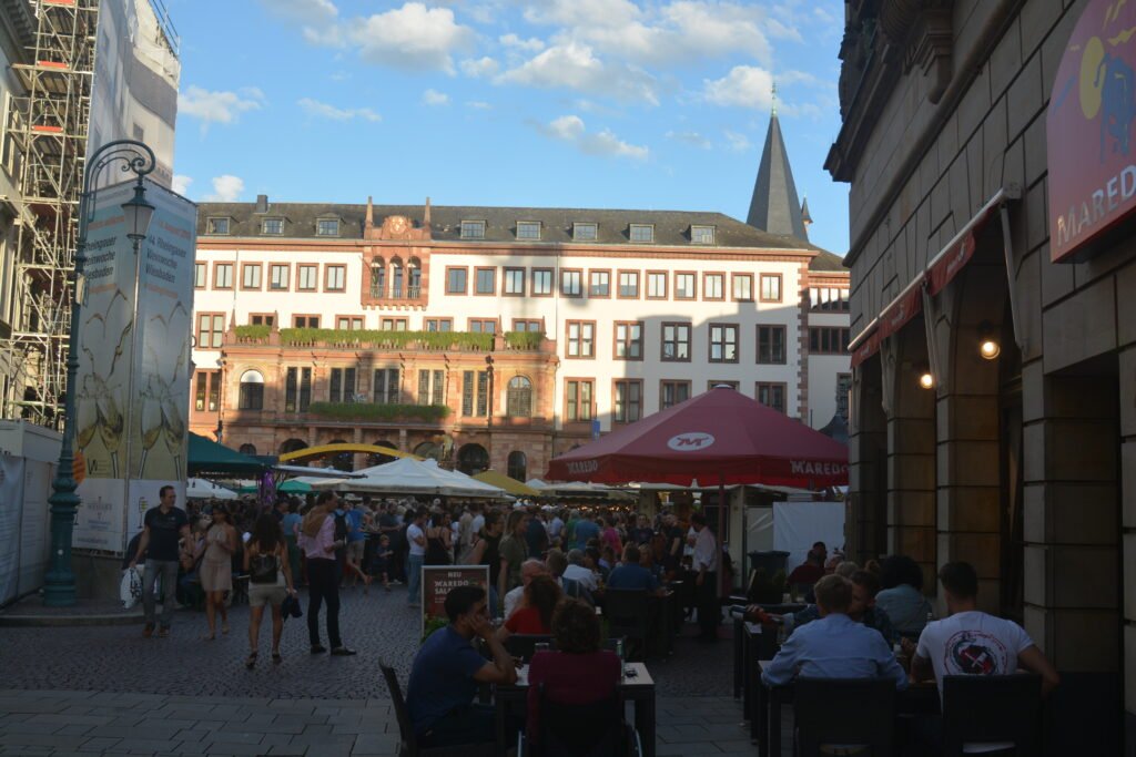 large open plaza surrounded by old and modern buildings. People are sitting a umbrella-covered tables in the plaza