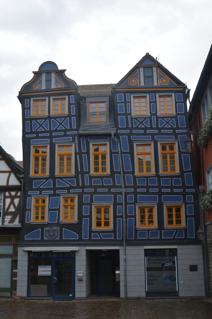 a four story half-timbered house painted blue with orange window frames, with a pronounced lean to the left