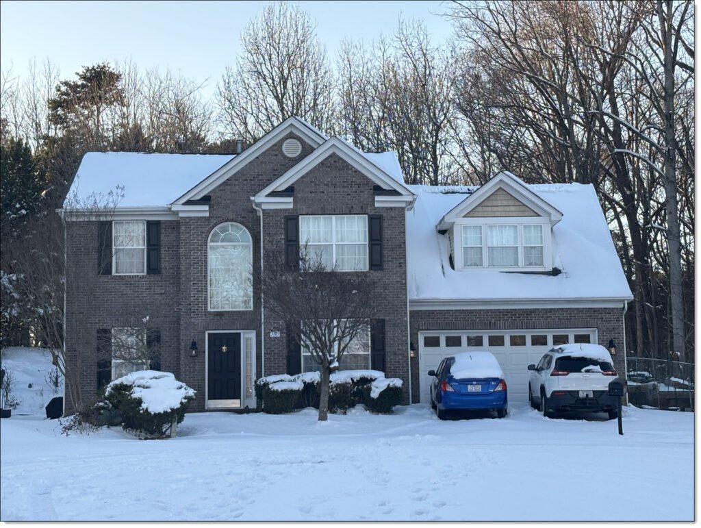 a house with roof covered in snow with a blue car and white car in the driveway. All the ground is blanked in snow.