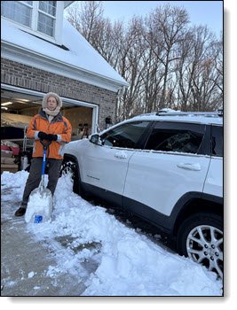 Ron wearing an orange coat and beige headcover, holding a snow shovel, standing in snow next to a white Jeep Cherokee.
