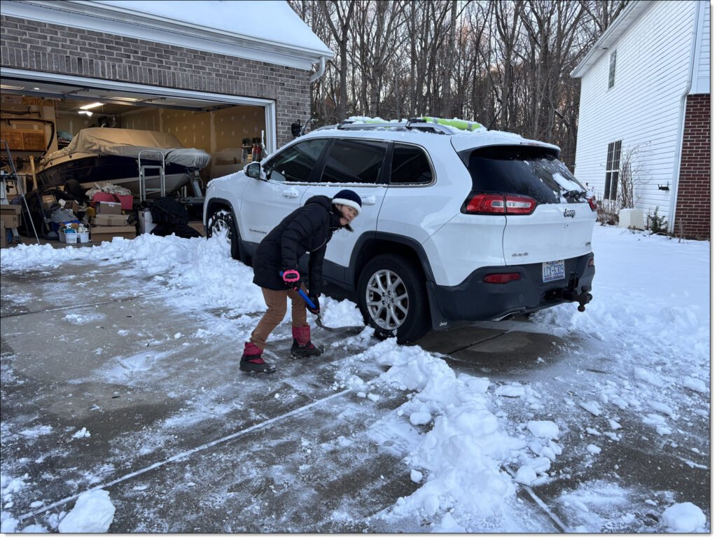 Winnie clearing snow from a large driveway with white Jeep Cherokee SUV next to her.