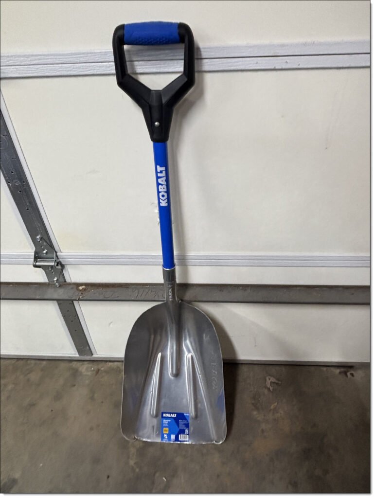 a shovel with wide aluminum scoop and blue shaft leaning against the inside of a garage door.