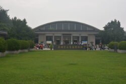 front view of a low-domed building with signs in chinese writing, sitting at the back of a large green lawn flanked by trees.
