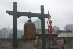 A Large Iron bell On The Ancient City Wall of Xi'an A large iron bell supported by a heavy wood frame, with red Chinese lanterns on the right side. A modern city skyline is in the background.