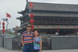 Ron and Weifang Charest standing in front of a chinese guardhouse with red chinese lanterns behind them.