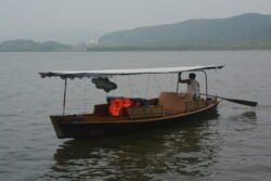 A Tourist Boat on West Lake a small wooden boat with a flat cloth roof. One person is in the back using a sculling oar to propel the boat.