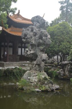 A Traditional Chinese Garden Along West Lake a jagged porous rock in the middle of a small pond in front of a Chinese pavilion with orange roof.