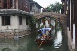 back view of a narrow canal boat being sculled under a stone arch bridge.