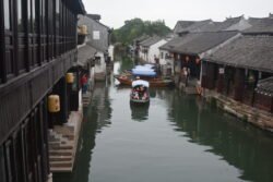 wide canal lined with stone buildings on each side. Many of the buildings have stone steps leading down to the water, and there are several sampan canal boats in teh canal.