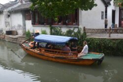 a small wooden boat with blue cloth awning roof, in a narrow canal with white building behind it, propelled by a man sitting in the back using a sculling oar.