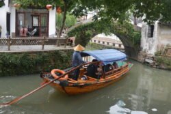 wooden sampan canala boat being sculled by a man standing up in back wearing a woven conical grass hat. The stone arch bridge is covered with green vines.