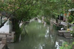 a waide canal with a canopy of trees, a pedestrian walkway on the right lined with shops and a canal boat in the distance.