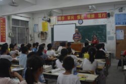 american man standing in front of a large class of chinese teenagers, being introduced by a Chinese woman. A green chalk blackboard in behind him and there are various red posters with chinese script above the blackboard. .
