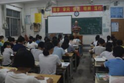 american man standing in front of a large class of chinese teenagers. A green chalk blackboard in behind him and there are various red posters with chinese script above the blackboard. .