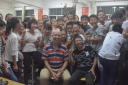 american man sitting in front of a large group of cheering chinese teenagers. Everyone is smiling and enjoying themselves.
