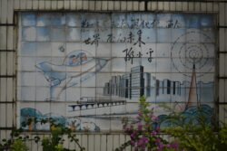Tile Mural On A Chinese School a tile mural with two people flying in a futuristic space plane, heading towards a city of clean hi-rise buildings with a modern train heading away.