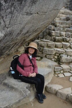 woman in red jacket with black packback and straw hat, sitting under a rock with stone steps behind her. taking