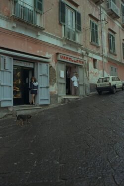 A young woman standing in the doorway of a Panificio next to a man standing in the doorway of a Macelleria, with shops on a steep hill and cobblestone roadway in front.
