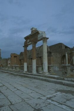 An ancient cobblestone road with three stone columns connected by a top stone with ruins of ancient building behind the columns