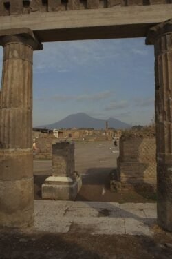 A view of an open site with broken columns, framed by two columns in the foreground. Mt Vesuvius is in the background.