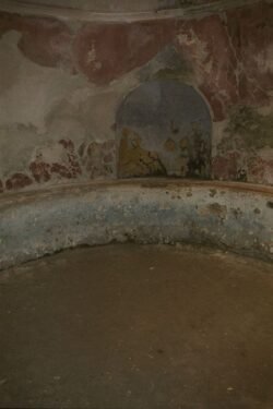 A curved fresco-covered stone wall with seating ledge and a deeper pool in the middle of the room.