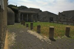 A walled courtyard partially covered in grass, partially covered with a gravel walkway. Open-faced building at the back built into the courtyard wall