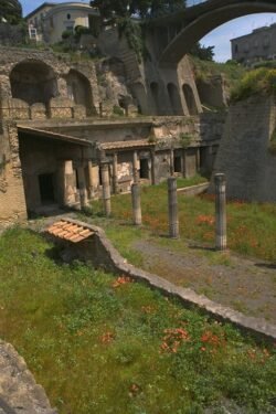 An ancient stone building tucked against a n earthen wall with columns lining a walkway to the front door. There is grass on one side of the walkway. Over the facade is an arched bridge structure.