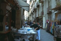 A street market on a narrow street surrounded by tall buildings with ornate facades.