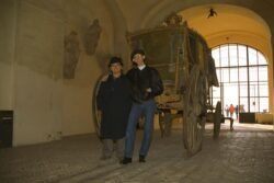 two people posing in front of an ornate horse-drawn carriage displayed in a museum
