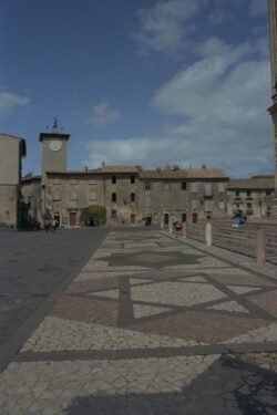 A large paved plaza in medieval style. A stone building with clock tower is at the back of the plaza.