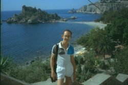 Ron wearing a multi-colored polo shirt and white shorts, standing on steps overlooking a beautiful coastal bay with a rocky island behind him.