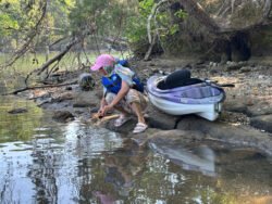 woman wearing a pink hat and blue lifejacket squatting along the edge of water, with a purple and white kayak pulled up on the lakeside beach.