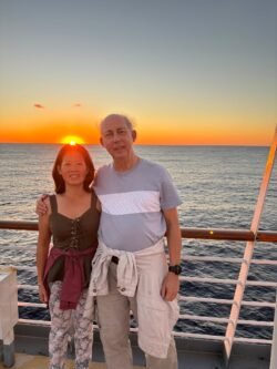 Ron and Winnie On A Cruise ship, against the rail, with sun rising behind them over the ocean.