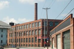 A large red brick factory building with numerous windows and a large cylindrical chimney extending above the building.