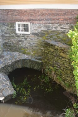 an ancient stone works of a wall with top of a tunnel over a small pool of water. A small four-pane glass window is mounted in the wall over the tunnel