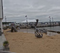 a large bronze sculpture of a disembodied man's left hand, right arm, bent knees, and a face coming out of a sand beach with water on the right side