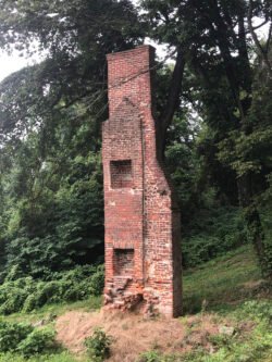 brick chimney with two recesses appearing to be fireplaces, extends like a tower over a grassy hill with tress behind it