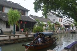 A sampan passing through a section of canal with wide plazas and step landings on the left side. Many people are on the plazas