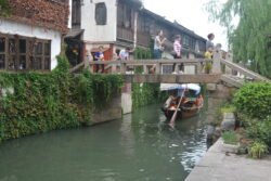 a flat-topped stone bridge with people walking across the top and a sampan canal boat passing underneath.
