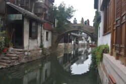 narrow canal lined with stone houses on each side and a stone arch bridge crossing the canal with people walking across. The bridge is reflected in the calm waters of the canal