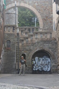 a man standing in front of an ancient stone wall with stairs on the left behind him, and an archway with door covered in modern graffiti