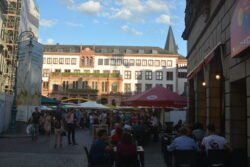 large open plaza surrounded by old and modern buildings. People are sitting a umbrella-covered tables in the plaza