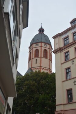 an octagon tower with domed roof and arched windows framed with red stone, viewed between buildings on either side.