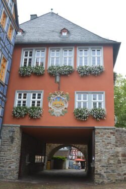 a red building with large double windows spanning a cobblestone road. One end of the building is supported on a thick stone wall