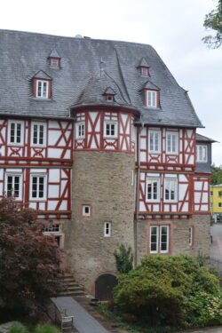 a large stone manor with a lower level of stone, upper levels of half-timbered construction, and a silo with conical roof on one side.