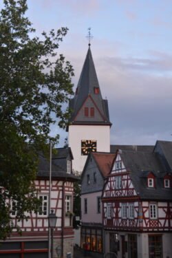 a church steeple with prominent clock face, Several half-timbered houses are below the steeple