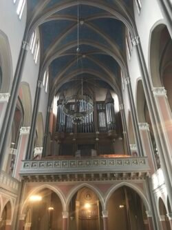 interior view of a cathedral with dramatic high vaulted ceiling, with the room ringed by tall stained glass windows and chandeliers.