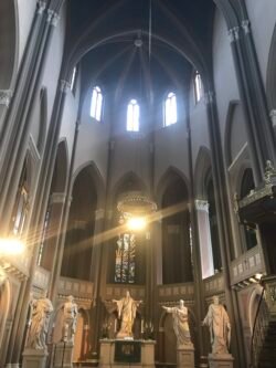 interior view of a cathedral with dramatic high vaulted ceiling, with the room ringed by tall stained glass windows and chandeliers. Along the floor level are several sculptures of religious figures.