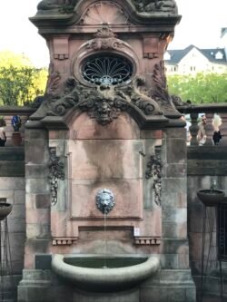 large decorative wall-mounted fountain with a bronze lions head streaming water into a basin built intot he fountain.