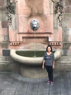 woman standing next to a stone fountain featuring a lion's head sculpture. water flows from the mouth of the detailed metal lion sculpture into a basin